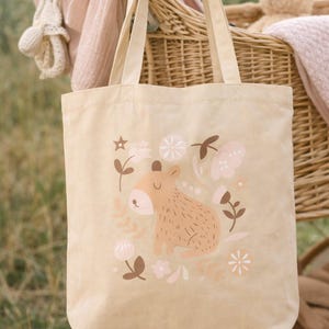 May include: Beige canvas tote bag featuring a whimsical illustration of a brown animal with floral accents in pink and brown. The bag has beige handles and is set against a backdrop of a wicker stroller and a teddy bear.