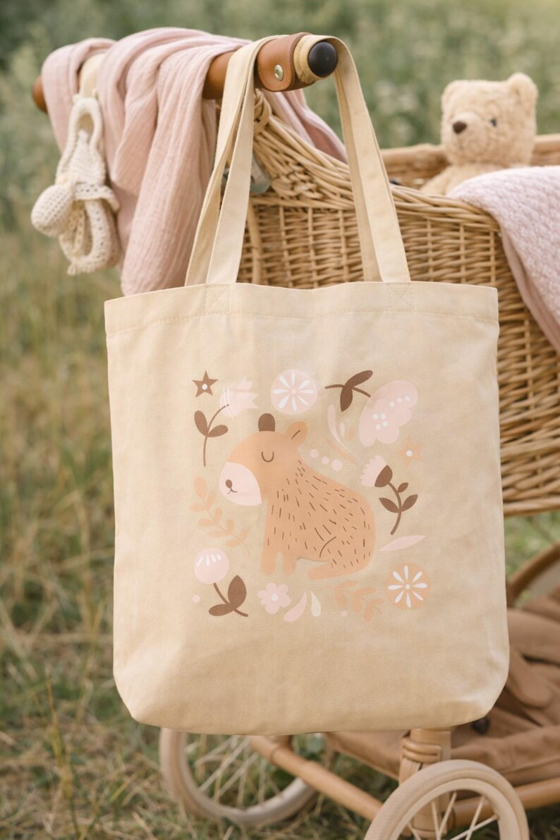 May include: Beige canvas tote bag featuring a whimsical illustration of a brown animal with floral accents in pink and brown. The bag has beige handles and is set against a backdrop of a wicker stroller and a teddy bear.