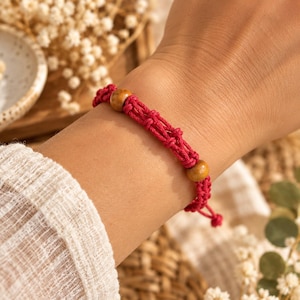 Peut inclure: Un bracelet en macramé rouge doté de deux perles en bois clair, lisses et rondes, intégrées dans le motif de corde nouée. Le bracelet est fabriqué à partir d'un cordon rouge vif noué avec des nœuds décoratifs, les perles étant espacées.