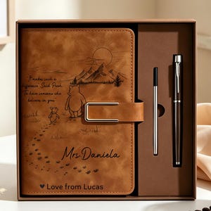 Könnte beinhalten: Ein hellbraunes Notizbuch in Lederoptik mit Metallverschluss, graviert mit einer Winnie Puuh-Illustration, Bergen und dem Text "It makes such a difference Said Pooh. To have someone who believes in you.", "Mrs. Daniela" und "Love from Lucas". Enthält einen passenden Stift.