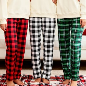 May include: Three pairs of pajama pants in red and black buffalo plaid, black and white checkered, and green plaid. The pajama pants are paired with cream-colored sweatshirts. The group is standing on a red and black plaid rug.