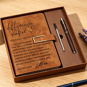 Könnte beinhalten: Ein braunes Notizbuch in Lederoptik in einer Geschenkbox mit passendem Stift und Mine. Der Einband zeigt den Text "difference maker" mit einem Herzsymbol, eine Definition im Wörterbuchstil und den Namen "Esther" in schwarzer Schreibschrift.
