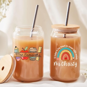 Könnte beinhalten: Zwei transparente Glasbecher mit Bambusdeckeln und Edelstahlstrohhalmen. Ein Becher hat ein Bleistift-, Apfel-, Kaffeetassen- und Lineal-Design mit dem Text "Mrs. Samantha". Der andere hat ein Regenbogen-Design und den Text "Ms. Christy".