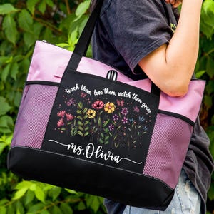 Peut inclure: Grand sac fourre-tout avec une base et des poignées noires, et un dessus rose. L'avant présente un panneau noir avec un motif floral et le texte "teach them, love them, watch them grow" et "ms. Olivia".