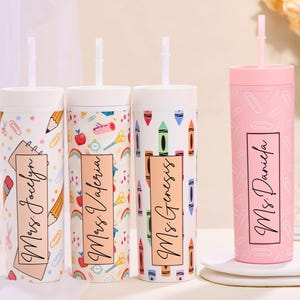 May include: Four personalized tumblers with straws. Three tumblers feature school-themed patterns with pencils, scissors, and crayons. The fourth tumbler is pink with a paperclip design. Each tumbler has a name in a rectangular frame.