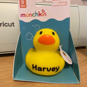 Puede incluir: Un patito de goma amarillo para el baño con pico naranja, que presenta el nombre Harvey impreso en negro en el pecho. El juguete está empaquetado en una caja de cartón con la etiqueta Munchkin White Hot Bath Ducky, 0 M+ y gráficos de burbujas.