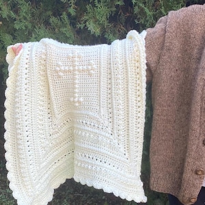 Puede incluir: Una manta de bebé de ganchillo blanco con un diseño de cruz en el centro. La manta tiene un borde festoneado y está hecha con un punto texturizado.