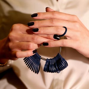 May include: A close-up of a person's hands holding a navy blue ring sizer tool. The person is wearing a navy blue ring and has black nail polish. The ring sizer tool is made of multiple navy blue rings attached to a metal bar.