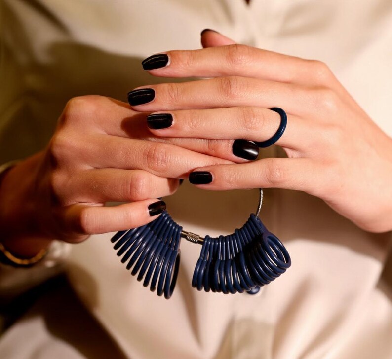 May include: A close-up of a person's hands holding a navy blue ring sizer tool. The person is wearing a navy blue ring and has black nail polish. The ring sizer tool is made of multiple navy blue rings attached to a metal bar.