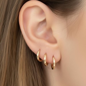 May include: Close-up of an ear with three gold hoop earrings of varying sizes. The earrings are simple, classic designs, reflecting a minimalist aesthetic. The ear is set against a backdrop of light brown hair and skin.