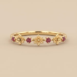 May include: A gold ring with a floral design, set with five ruby gemstones. The ring features three flower-shaped settings and two round ruby settings. The ring is displayed against a neutral background.