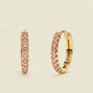 May include: A pair of gold hoop earrings, each encrusted with small pink gemstones. The earrings are displayed against a soft, neutral background, highlighting their color and design. The gemstones are evenly spaced around the hoops.