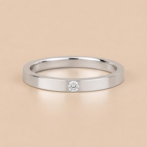 May include: A polished silver-toned metal ring featuring a flat band design with a single, small, round, clear faceted gemstone set flush into the centre of the band, creating a smooth and continuous surface profile for the jewellery piece.