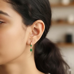 Puede incluir: Pendientes de aro dorados con un diseño floral verde colgante. Los pendientes presentan un pequeño aro dorado que se conecta a un grupo de piedras preciosas verdes en forma de flor. Los pendientes se muestran en la oreja de una persona.