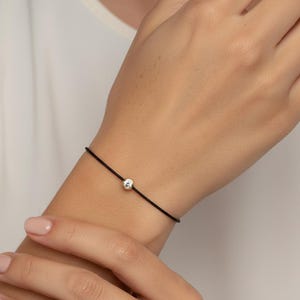 Puede incluir: Una pulsera minimalista de cordón negro con una sola gema redonda y transparente. La gema está engastada en un engaste de plata u oro blanco. La pulsera se lleva en la muñeca.