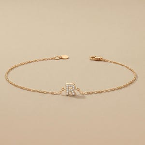 Pode incluir: Uma delicada pulseira de corrente dourada com a letra "R" adornada com pequenas pedras brilhantes. A pulseira tem um fecho simples e um pequeno pingente redondo. A pulseira é exibida em um fundo neutro.