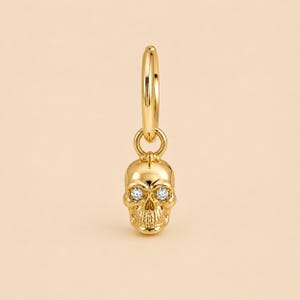 Puede incluir: Un pendiente de aro dorado con un dije de calavera colgante. La calavera está hecha de metal pulido en tono dorado y presenta dos pequeñas piedras preciosas redondas y transparentes engastadas en las cuencas de los ojos, aportando brillo.