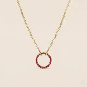 May include: A gold-colored necklace with a circular pendant. The pendant is encrusted with small, red gemstones. The necklace chain is delicate and hangs from the pendant. The background is a solid, light beige color.