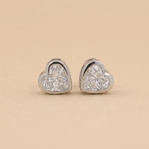 May include: A pair of heart-shaped stud earrings. Each earring features a clear, faceted gemstone set in a silver-colored metal bezel. The earrings are displayed on a neutral, light-colored background.