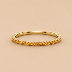 Op de afbeelding: Een dunne gouden ring met een doorlopende rij kleine, ronde, gefacetteerde gele citrien edelstenen die langs de gehele omtrek zijn gezet. Het metaal heeft een gepolijste afwerking en de stenen zijn uniform in grootte en kleur over het hele sieraad.
