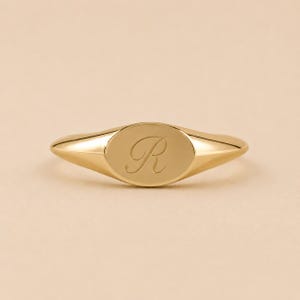May include: A gold signet ring featuring an oval face with a polished finish. The center of the oval face is engraved with the capital letter R in a cursive script font. The band of the ring tapers smoothly from the oval face to the back.