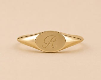 Puede incluir: Un anillo de sello de oro pulido con una cara lisa de forma ovalada. El centro del óvalo está grabado con una sola letra R elegante en cursiva. La banda se estrecha desde el óvalo central, creando una forma metálica elegante y continua.