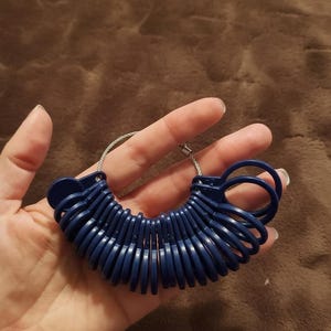 May include: A set of navy blue ring sizers on a silver wire loop. The ring sizers are arranged in a semi-circle, held in a hand against a brown background. The sizers are made of plastic and are used to measure ring sizes.