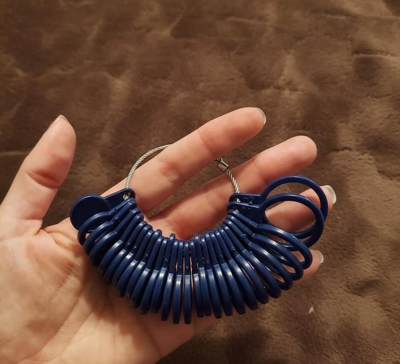 May include: A set of navy blue ring sizers on a silver wire loop. The ring sizers are arranged in a semi-circle, held in a hand against a brown background. The sizers are made of plastic and are used to measure ring sizes.