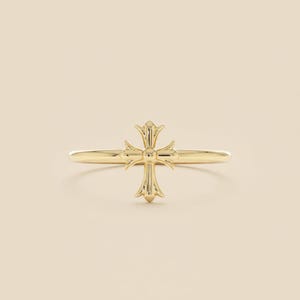 May include: A gold-colored ring featuring a detailed cross design. The cross is centered on the ring's band, with ornate detailing on each arm. The ring has a polished finish, reflecting light. The background is a neutral, solid color.
