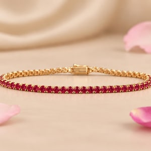 May include: A gold tennis bracelet featuring a continuous row of round, vibrant red gemstones set in a polished gold metal frame, complete with a secure rectangular box clasp mechanism visible at the center of the circular arrangement.