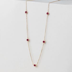 Puede incluir: Un delicado collar de oro con cinco piedras preciosas redondas de color rojo intenso, espaciadas uniformemente a lo largo de la cadena. El collar se muestra sobre un fondo blanco limpio, resaltando la elegancia sencilla de la joya y el color vibrante de las piedras.