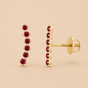 May include: A pair of gold ear climbers featuring six round-cut red rubies set in a curved vertical line. The earrings include a threaded post and a circular screw-back closure mechanism, showcasing a polished gold finish throughout the metal setting.