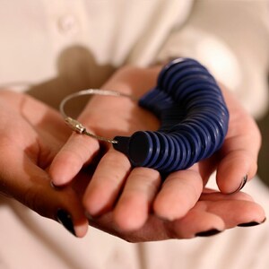 May include: A blue ring sizer tool with multiple ring sizes attached to a silver metal wire. The ring sizer is held in open hands against a white background. The ring sizer is a tool used to measure finger sizes for rings.