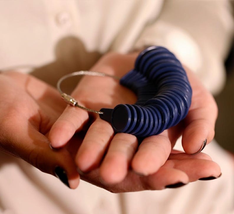 May include: A blue ring sizer tool with multiple ring sizes attached to a silver metal wire. The ring sizer is held in open hands against a white background. The ring sizer is a tool used to measure finger sizes for rings.