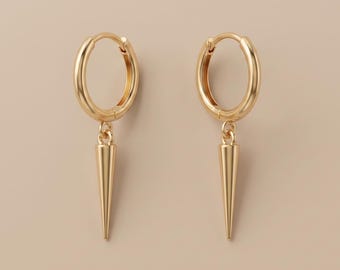 May include: A pair of gold-colored hoop earrings. Each earring features a small, oval hoop with a dangling, cone-shaped charm. The earrings are displayed against a neutral, light brown background.