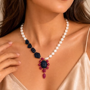 May include: A necklace featuring a strand of white pearls transitioning into three dark blue textured geometric beads. The centerpiece is a large oval dark blue textured stone surrounded by small magenta textured beads in a floral arrangement.