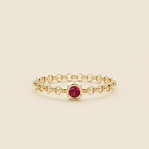 Puede incluir: Un anillo dorado con un diseño de eslabones de cadena. El anillo presenta una gema roja redonda en el centro. El anillo está sobre un fondo crema, resaltando los detalles de la joya.