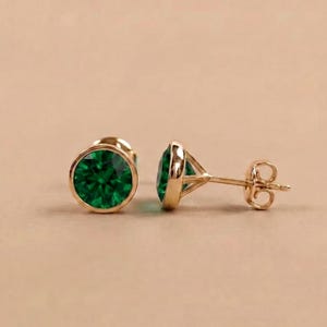 May include: A pair of gold stud earrings with round, vibrant green gemstones. The gemstones are set in a bezel setting, and the earrings have a classic, elegant design. The earrings are displayed on a neutral background.