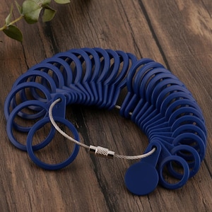 May include: A set of blue plastic ring sizers connected by a silver-colored wire. The ring sizers are arranged in a semi-circle, displaying various ring sizes. The background is a dark wooden surface.