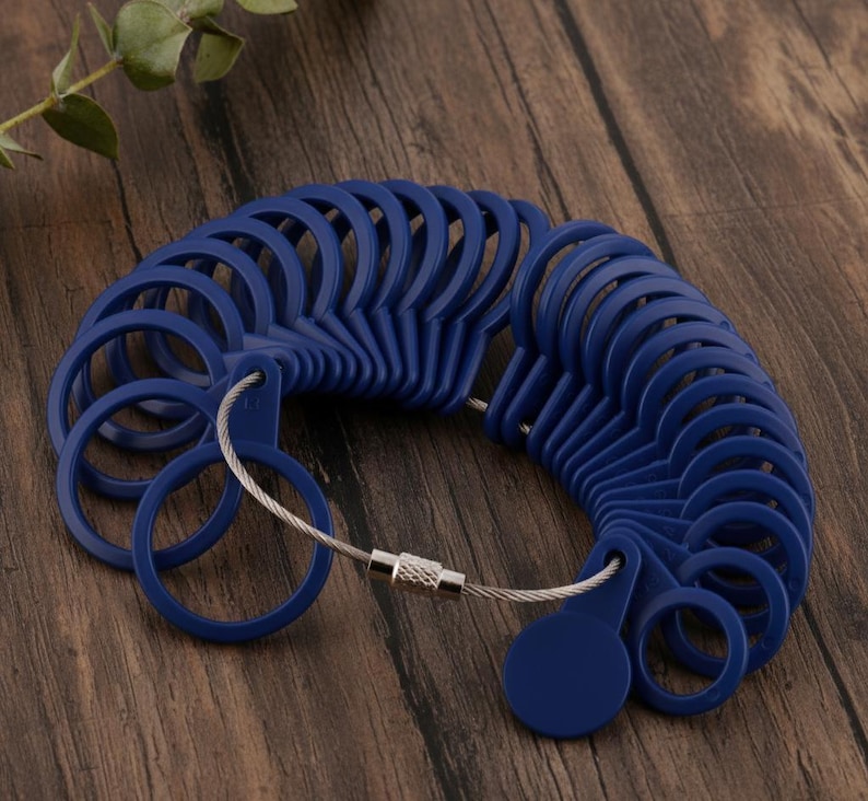 May include: A set of blue plastic ring sizers connected by a silver-colored wire. The ring sizers are arranged in a semi-circle, displaying various ring sizes. The background is a dark wooden surface.