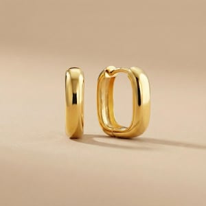 May include: Two gold-colored, rectangular hoop earrings. The earrings have a smooth, polished surface and a rounded rectangular shape. The earrings are displayed on a neutral background.