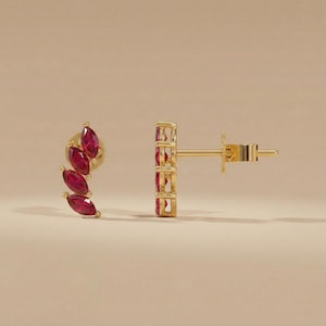 May include: A pair of gold-colored stud earrings. Each earring features three marquise-cut, ruby-red gemstones. The earrings are set in a gold-colored metal and have a post and butterfly backing.