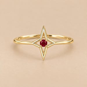 May include: A gold ring featuring a four-pointed star design centered with a small, round, deep red gemstone. The band is thin and polished, with the star motif extending vertically to create a symmetrical, geometric focal point on the finger.