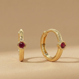 以下が含まれることがあります： 各イヤリングの中央に小さな丸い赤い宝石がセットされた、2つの金のフープイヤリング。イヤリングは、柔らかい照明を備えた、ニュートラルな明るい茶色の表面に表示されています。