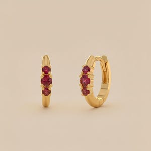 May include: A pair of gold hoop earrings, each adorned with three vibrant red gemstones. The earrings have a polished finish, reflecting light. The gemstones are evenly spaced along the front of the hoops, creating a classic and elegant look.