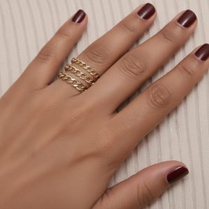 Puede incluir: Un conjunto de tres anillos de metal en tono dorado, cada uno con un diseño clásico de cadena de eslabones. Los anillos están apilados en un dedo, mostrando un acabado metálico pulido y eslabones entrelazados uniformes.