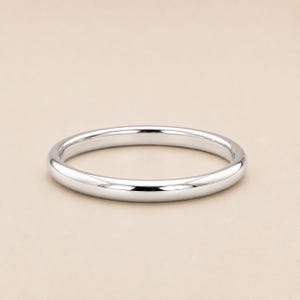Puede incluir: Un anillo de metal plateado pulido con una banda lisa y redondeada. El anillo presenta un diseño circular simple y continuo con una superficie reflectante que capta la luz, creando reflejos sutiles a lo largo de los bordes curvos de la banda.