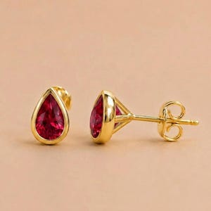 Puede incluir: Un par de pendientes de botón de oro con piedras preciosas de color rojo intenso en forma de pera, engastadas en un bisel de oro liso. Los pendientes incluyen un poste y un cierre de mariposa de oro a juego, con un acabado metálico pulido.