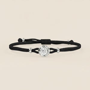 Puede incluir: Pulsera de cordón negro con un elemento central plateado en forma de flor con una gema redonda y transparente. La pulsera incluye pequeñas cuentas plateadas y un cierre de nudo ajustable.