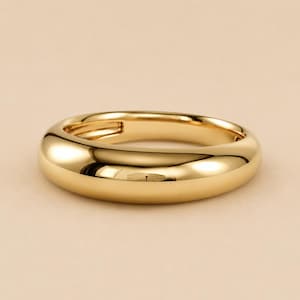 May include: A polished gold ring with a thick, rounded band and a smooth, reflective surface. The jewellery piece features a continuous, solid metal construction with a high-shine finish that catches the light, creating bright highlights.